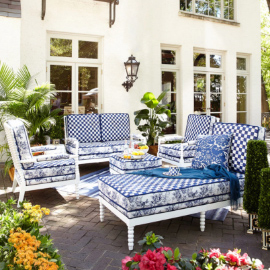 Blue and white patio furniture with a snack tray in a backyard, surrounded by colorful flowers and greenery, behind a white house