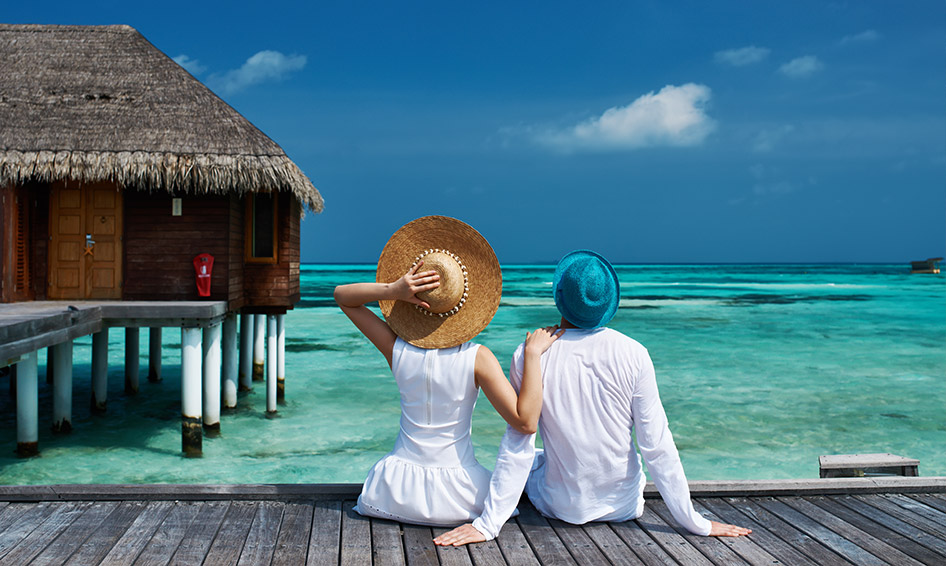 Wooden lounge chairs lined up in a row on a beach near a palm tree and across the water from a group of thatch roof bungalows. The text A Honeymoon In Paradise is written beneath on a mint green background. 