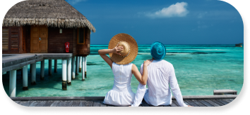 Young couple dressed in white sit on dock looking out onto clear blue ocean with a hut jetting out into the water to the left of couple.